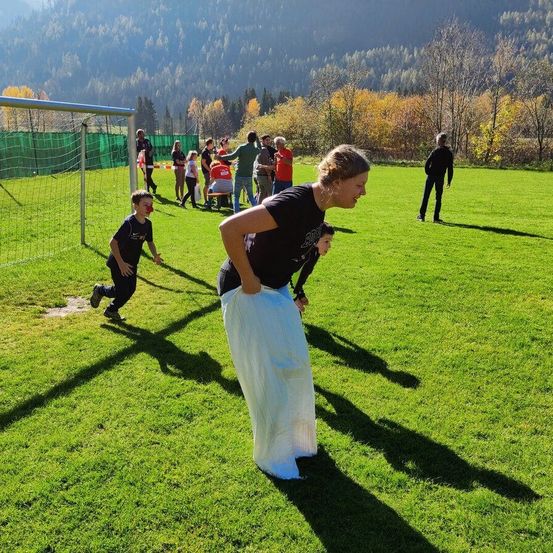 Eine Frau rennt auf einen Jungen auf einem Rasenfeld zu. In der Nähe stehen ein Fußballtor und eine Gruppe von Menschen, die stehen und sitzen. Bäume und Berge sind im Hintergrund.
