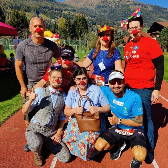 Eine Gruppe von Personen in spielerischen Kostümen, einschließlich roter Clownsnasen, posiert auf einer Laufbahn mit Bergen im Hintergrund.