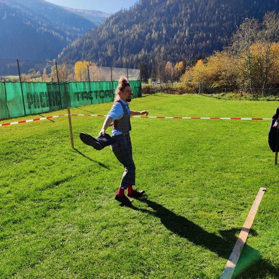 Ein Mann in einem karierten Outfit spielt ein Spiel auf einem Feld mit Bergen im Hintergrund.