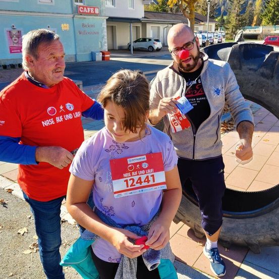 Drei Personen stehen in der Nähe eines Gebäudes, ein Mann hält einen Ballon und ein Mädchen trägt einen Nummernaufkleber mit der Nummer 12441.