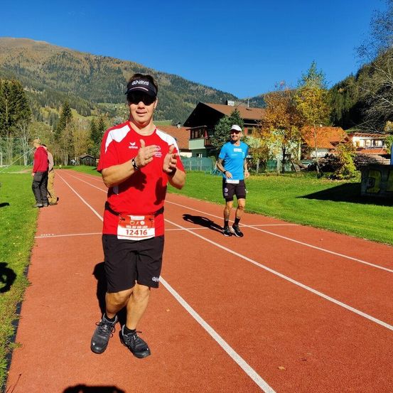 Ein Mann in einem roten Shirt steht auf einer roten Laufbahn mit Bergen im Hintergrund. Er trägt eine Sonnenbrille und eine Uhr. Hinter ihm läuft ein weiterer Mann.