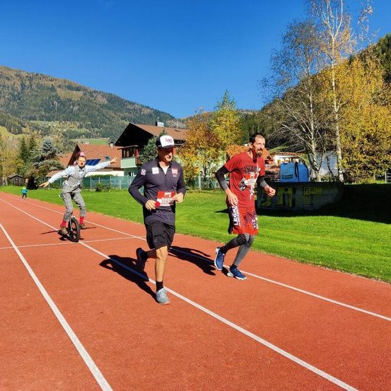 Drei Männer laufen auf einer roten Laufbahn mit weißen Linien. Der Mann vorne trägt eine Kappe und Turnschuhe. Die anderen beiden tragen rote Hemden und Turnschuhe. Bäume und Berge sind im Hintergrund.