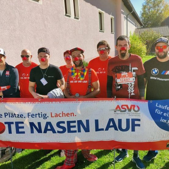 Eine Gruppe von Menschen mit roten Nasen und Clown-Kostümen steht hinter einem Banner. Das Banner trägt die Aufschrift 'TE NASEN LAUF'. Sie sind draußen vor einem Gebäude.