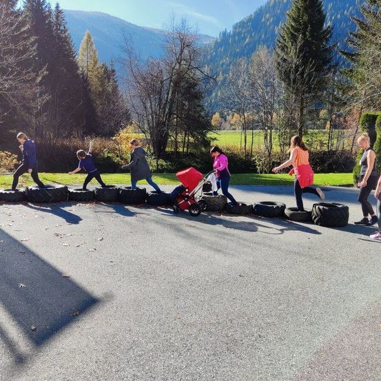 Mehrere Menschen laufen auf Reifen auf der Straße. Eine Frau mit einem Kinderwagen ist in der Mitte. Der Boden ist mit gefallenen Blättern bedeckt. Es gibt Bäume und Berge im Hintergrund.