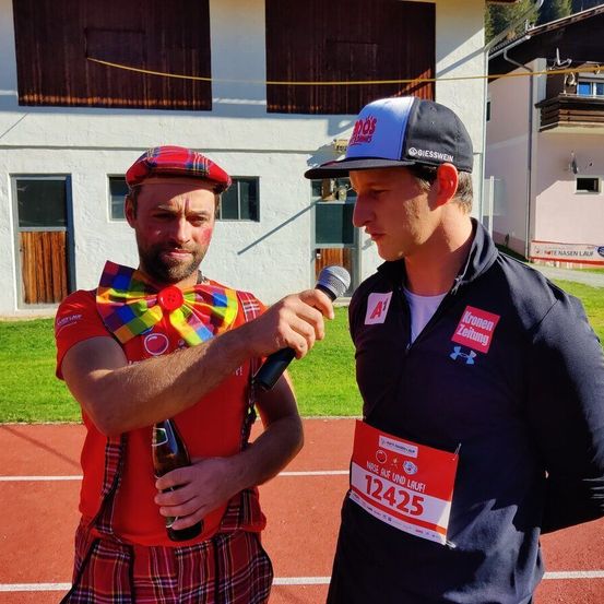 Ein Mann in einem roten Clownskostüm wird von einem anderen Mann in einer schwarzen und weißen Baseballkappe interviewt. Er hält ein Mikrofon und eine Flasche. Der Interviewte trägt ein rotes Namensschild mit der Nummer 12425.