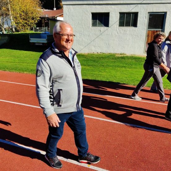 Ein Mann in einem grauen Jacke steht lächelnd auf einer Laufbahn. Hinter ihm gehen zwei Frauen auf der Bahn. Ein Gebäude mit einem Lastwagen in der Nähe ist im Hintergrund.