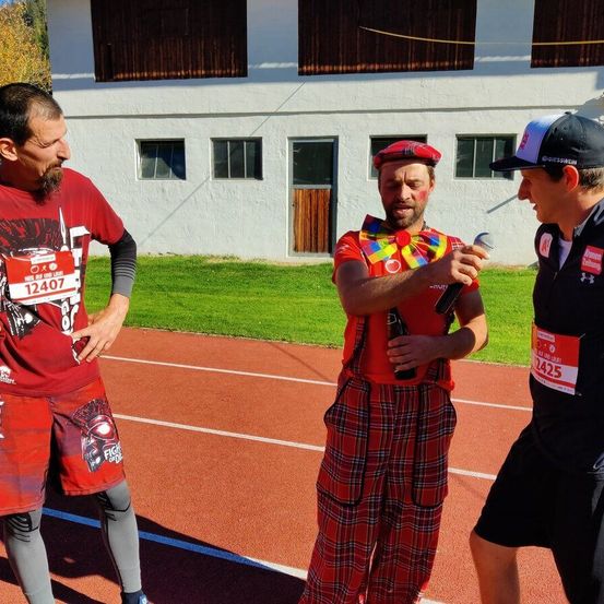 Drei Männer auf einer Laufbahn, einer hält ein Mikrofon, wahrscheinlich interviewt er die anderen. Der Mann in der Mitte trägt einen roten Hut und eine bunte Weste.