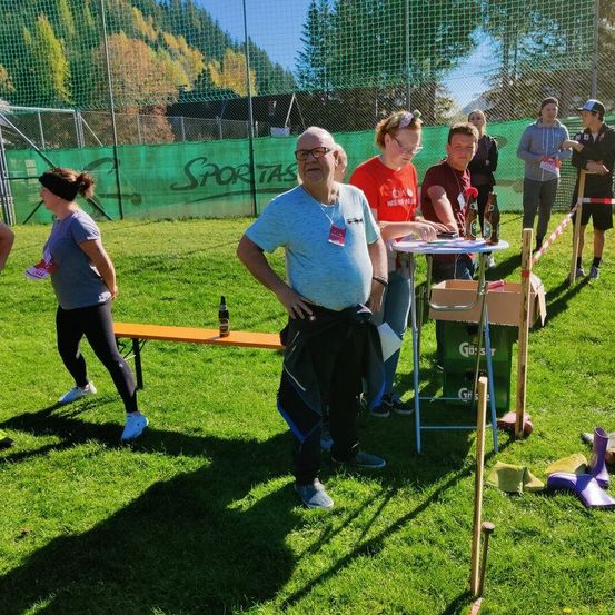 Eine Gruppe von Menschen versammelt sich im Freien, wobei ein Mann in der Nähe eines Tisches steht. Einige Personen gehen in der Nähe. Die Umgebung umfasst eine Bank, Flaschen und ein Netz im Hintergrund.