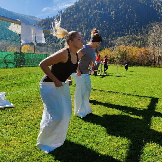 Zwei Frauen lachen, während sie weiße Säcke tragen, auf einem Rasen. Sie stehen neben einem Fußballtor mit Bergen im Hintergrund.
