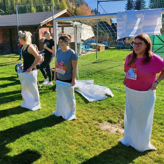 Vier Frauen stehen auf einem Rasenfeld in weißen Säcken, möglicherweise an einem Sackrennen teilnehmend. Sie stehen neben einem Fußballtor mit Netz. Dahinter befindet sich ein Gebäude, und Autos sind davor geparkt.