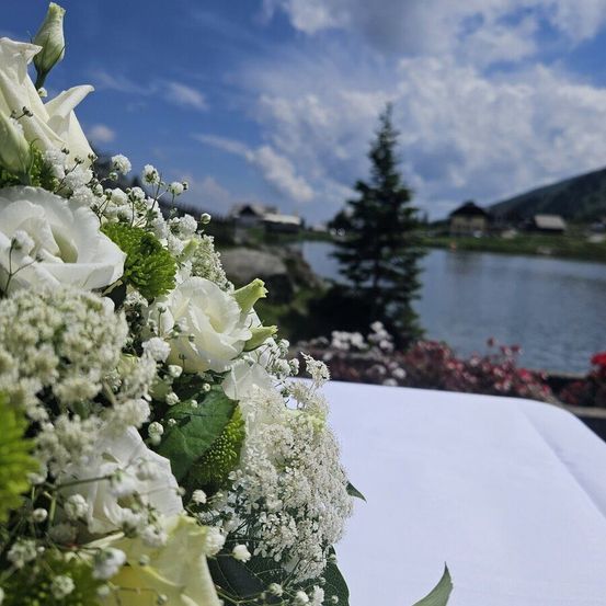 Ein Hochzeitsstrauß aus weißen Blumen steht auf einem Tisch in der Nähe eines Sees mit Bergen im Hintergrund.