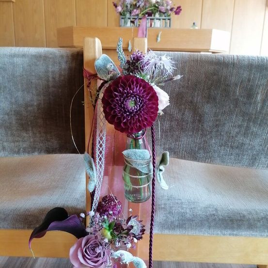 Ein hölzerner Gangstuhl, geschmückt mit einem Strauß lila Blumen und einem klaren Einmachglas. Der Stuhl ist von anderen Holzstühlen umgeben.