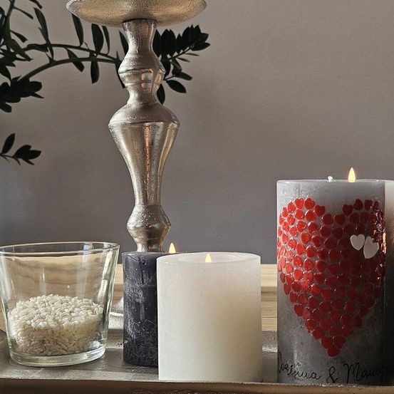Auf einem Tisch sind ein silberner Kerzenständer, eine Kerze mit Reis, eine schwarze Kerze und eine weiße Kerze mit Herzdesign zu sehen. Der Hintergrund ist grau.