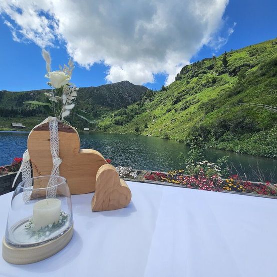 Eine ruhige Umgebung mit einem See und Bergen im Hintergrund. Eine Kerze befindet sich in einer Glasschale, und ein hölzerne Herz mit einer weißen Rose ist in der Nähe.