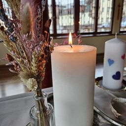 Zwei weiße Kerzen auf einem Tisch, eine entzündet. Eine Vase mit getrockneten Blumen neben der entzündeten Kerze. Eine weitere Kerze mit Herzdesigns auf einem Teller. Ein Fenster mit Blick auf Gebäude draußen.