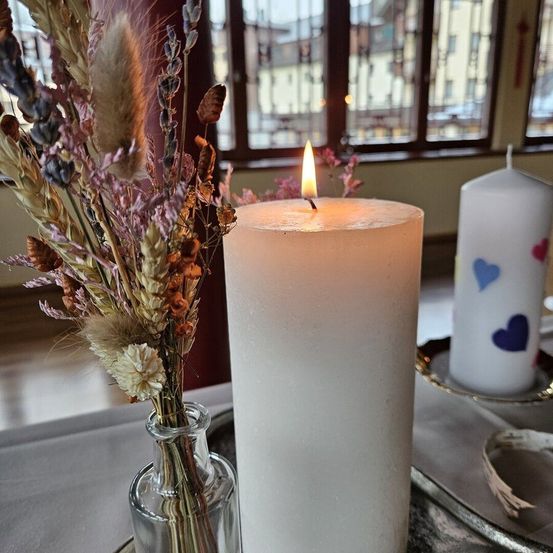 Zwei weiße Kerzen auf einem Tisch, eine entzündet. Eine Vase mit getrockneten Blumen neben der entzündeten Kerze. Eine weitere Kerze mit Herzdesigns auf einem Teller. Ein Fenster mit Blick auf Gebäude draußen.