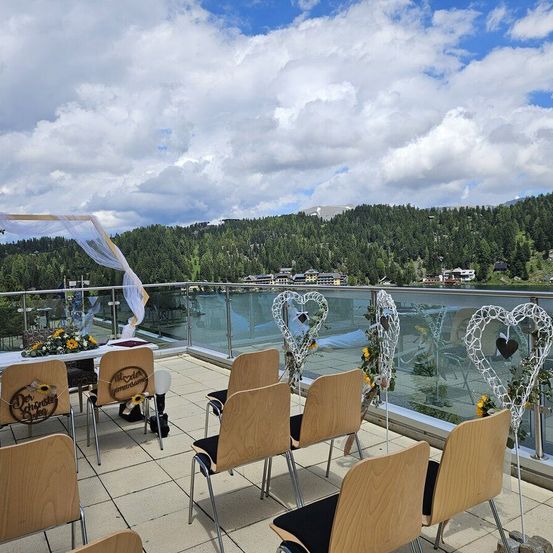 Eine Hochzeitszeremonie auf einer Dachterrasse mit Blick auf Berge, mit einem Tisch mit Blumen, Holzstühlen und dekorativen herzförmigen Rahmen.