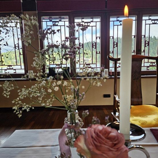 Ein Tisch ist mit einer Kerze und einer Blumenvase in einem Raum mit gemusterten Fenstern gedeckt. Ein Stuhl steht in der Nähe.