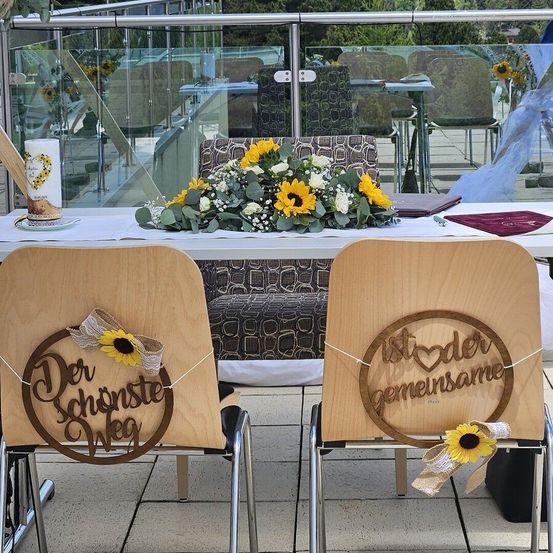 Zwei Stühle an einem Tisch mit Sonnenblumen und einem deutschen Text, 'Der schonste Weg' und 'Wirst der gemeinsamsame.' Ein Blumenstrauß steht auf dem Tisch, und eine Glaswand befindet sich dahinter.