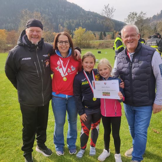 Eine Familie steht auf einem Rasenfeld mit einem Berg im Hintergrund. Zwei Mädchen mit Medaillen und einem Zertifikat stehen zwischen zwei Männern. Sie alle lächeln.