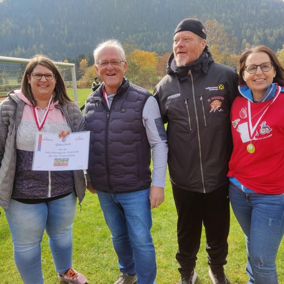 Vier Personen stehen auf einem Rasenfeld mit einem Fußballtor im Hintergrund. Sie tragen Sporttrikots und Medaillen, und eine von ihnen hält ein Zertifikat.