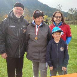 Eine vierköpfige Familie steht auf einem Rasen und lächelt. Der Mann trägt eine schwarze Jacke, die Frau eine graue Jacke und der Junge einen blauen Hoodie. Sie tragen Medaillen, was darauf hindeutet, dass sie an einer Veranstaltung teilgenommen haben. Hinter ihnen ist ein Berg sichtbar.