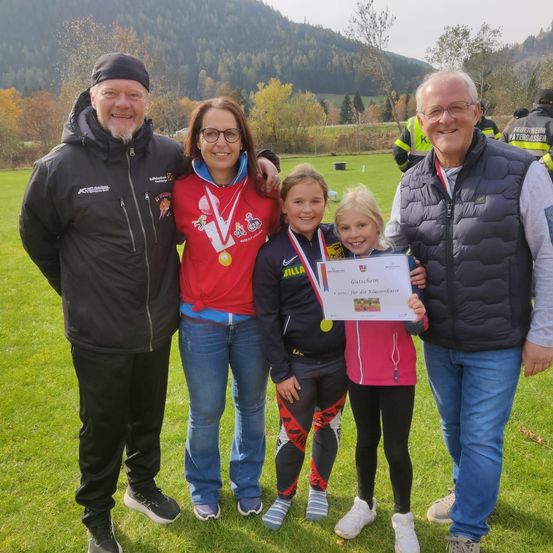 Eine Familie mit zwei Mädchen und zwei Männern steht auf einem Grasfeld und posiert für ein Foto. Sie tragen Sportjacken, Hosen und Turnschuhe. Sie haben Medaillen und ein Zertifikat. Dahinter ist ein verschwommener Blick auf einen Berg.