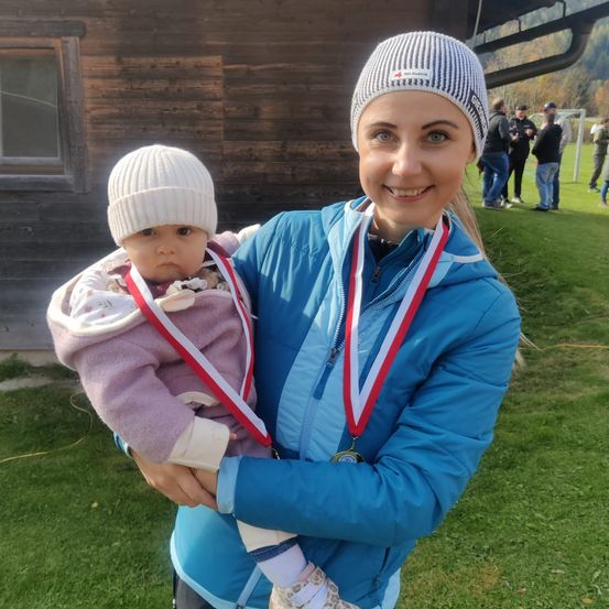 Eine Frau in einer blauen Jacke hält ein Baby, das Medaillen trägt. Beide tragen Mützen. Dahinter steht ein Gebäude und ein Grasfeld mit stehenden Personen.