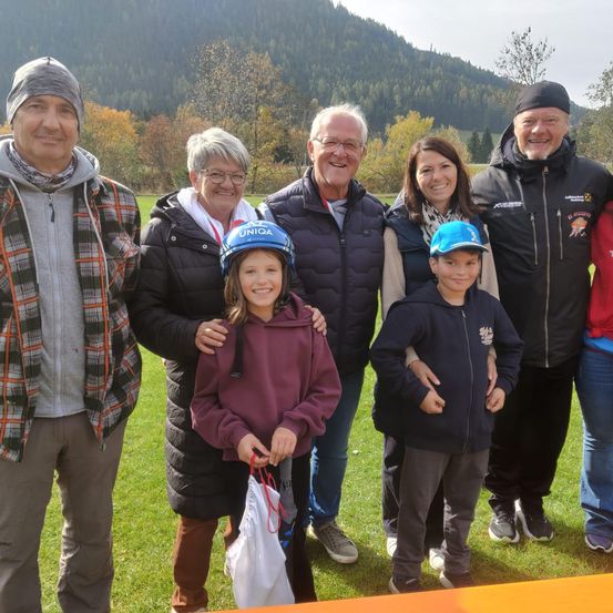 Eine Gruppe von Erwachsenen und Kindern posiert für ein Foto auf einem Rasenfeld mit einem Berg im Hintergrund. Zwei Kinder tragen Helme.