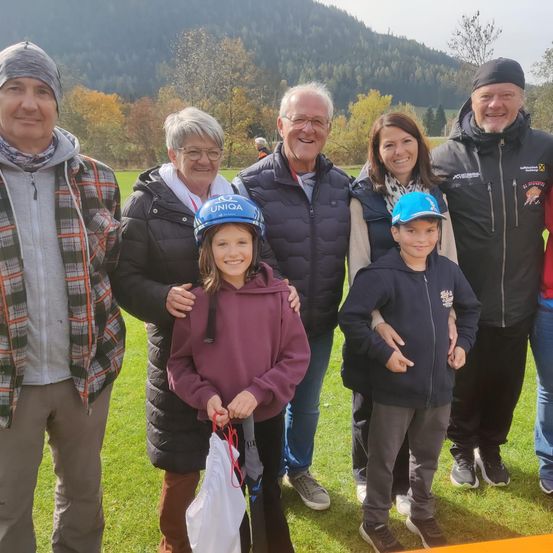 Eine sechsköpfige Familie steht lächelnd auf einem Rasenfeld. Sie tragen Jacken und Helme. Hinter ihnen befindet sich ein Berg mit Bäumen.
