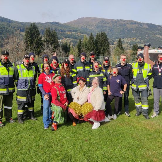 Eine Gruppe von Feuerwehrleuten mit roten Clownsnasen posiert für ein Foto auf einer Wiese mit Bergen im Hintergrund.