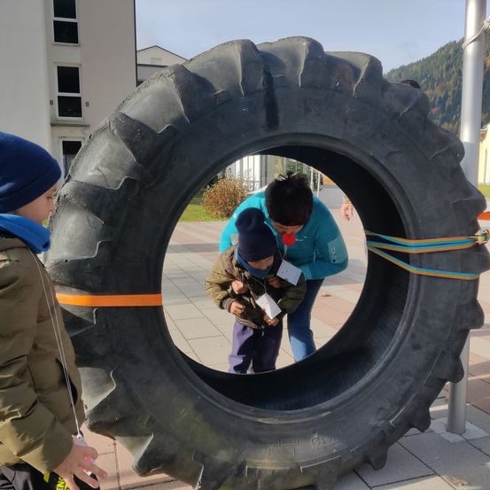 Drei Kinder erkunden einen großen Reifen, wobei eines hineinschaut. Sie befinden sich auf einer gepflasterten Fläche mit Gebäuden im Hintergrund.