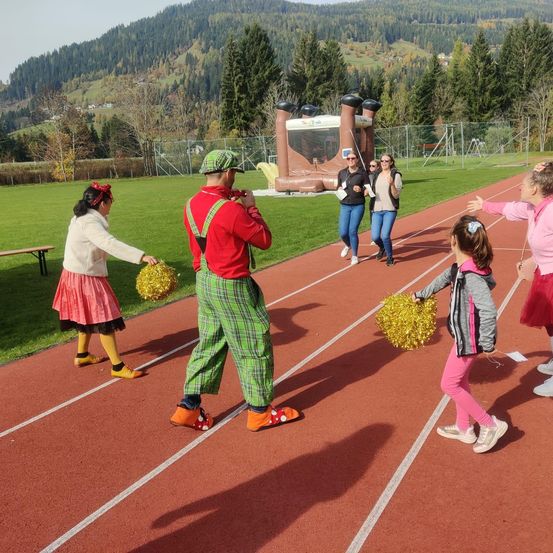 Eine Gruppe von Menschen läuft auf einer Laufbahn, darunter ein Mann in Clownskostüm und eine Frau mit Pom-Pom. Sie sind von Bäumen und Bergen im Hintergrund umgeben.