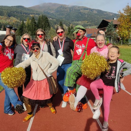 Eine Gruppe von Menschen in Kostümen, darunter Clowns, posiert für ein Foto auf einer Laufbahn mit Bergen im Hintergrund.