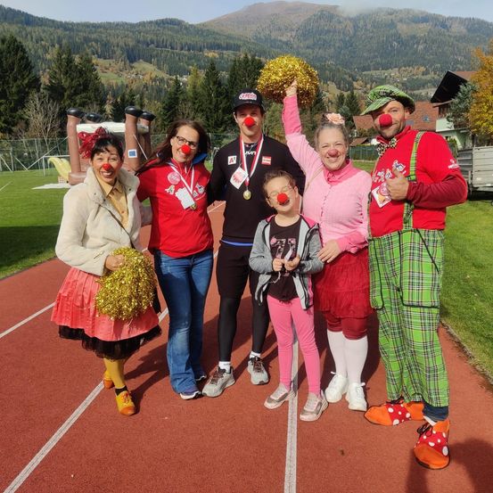 Eine Gruppe von Menschen in Clownkostümen und mit Pom-Poms posiert für ein Foto auf einer Laufbahn mit Bergen im Hintergrund.