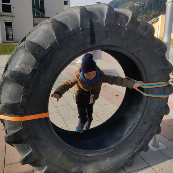 Ein junges Kind in einem blauen Hut und einer braunen Jacke kriecht durch einen großen Reifen auf einem Ziegelweg. Hinter dem Reifen befindet sich ein Gebäude mit Glasfenstern und einem geneigten Bereich mit Bäumen.