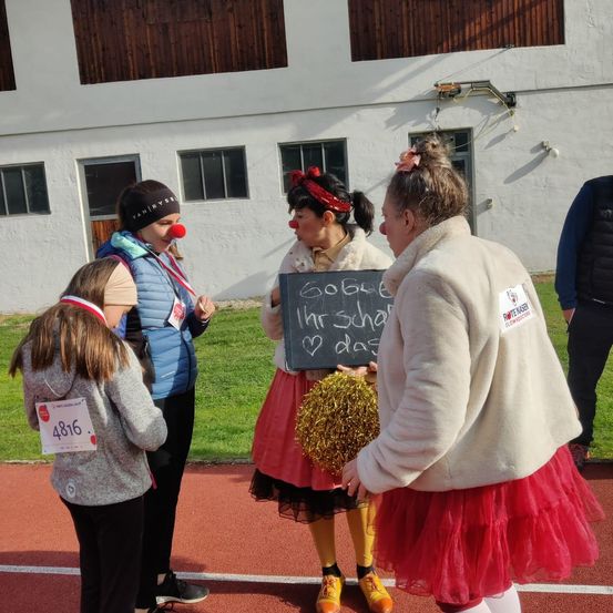 Eine Gruppe von Menschen, einige in Kostümen, stehen auf einer Laufbahn mit einem Gebäude im Hintergrund. Eine Person hält ein Schild mit der Aufschrift 'Golle Ihr Schatz' und eine andere hat einen Pom-Pom.
