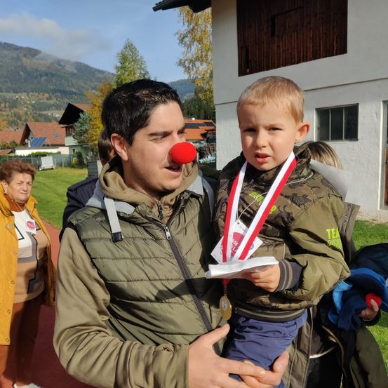 Ein Mann mit einer roten Clownsnase hält einen jungen Jungen mit Medaillen um den Hals auf einer Rasenfläche.
