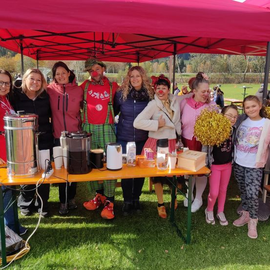 Eine Gruppe von Menschen, darunter zwei Clowns, steht unter einem roten Zelt bei einer Outdoor-Veranstaltung, lächelt und posiert für ein Foto. Sie befinden sich in der Nähe eines Tisches mit verschiedenen Gegenständen, wie einem Wasserkocher und Tassen.