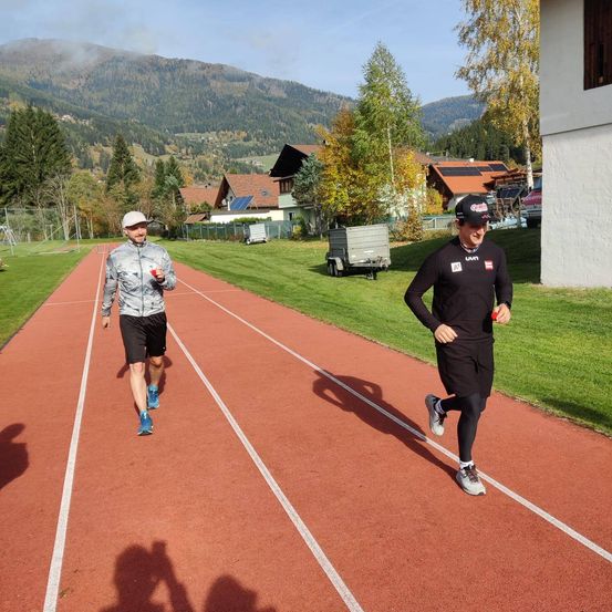 Zwei Männer joggen auf einer roten Laufbahn, einer hält einen roten Gegenstand in der Hand, in der Nähe von Häusern mit Solarpanelen und Bergen im Hintergrund.