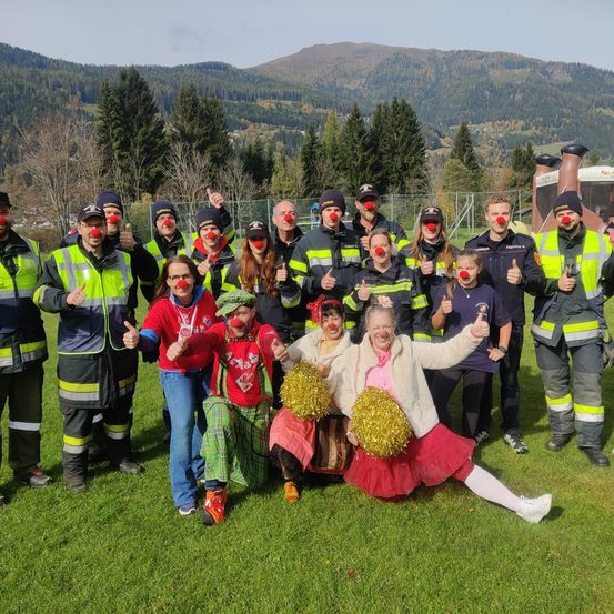 Eine Gruppe von Feuerwehrleuten in Clownskostümen posiert für ein Foto auf einer Rasenfläche mit Bergen im Hintergrund.