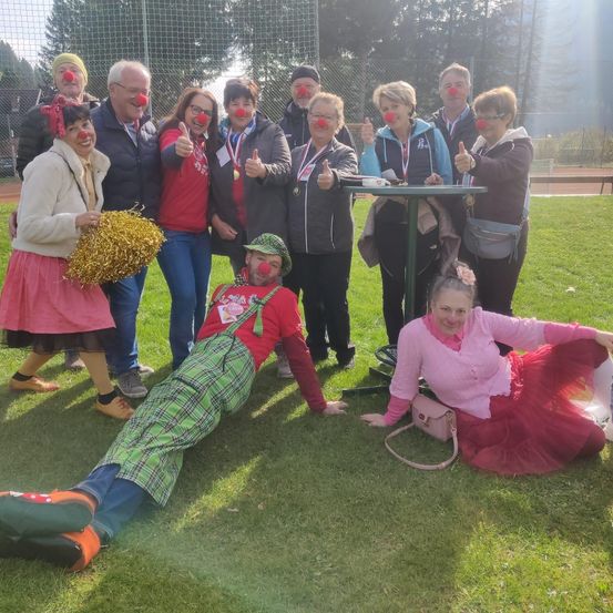 Eine Gruppe von Menschen in Clownkostümen posiert für ein Foto auf einem Rasenfeld, einige halten eine Trophäe und einen Tisch im Hintergrund.