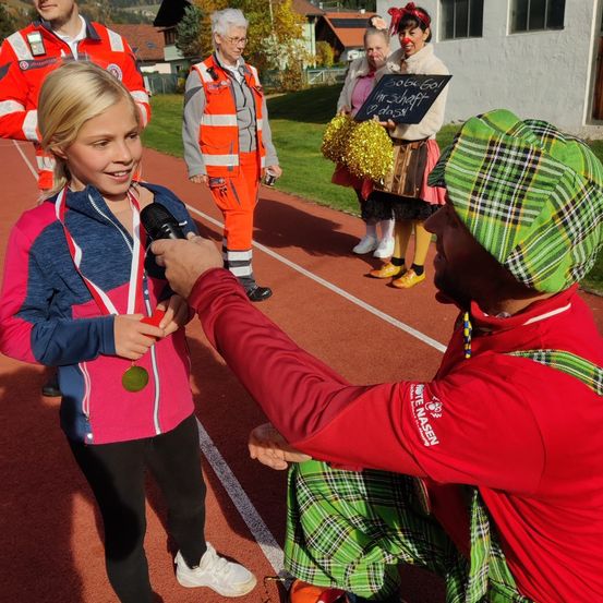 Ein junges Mädchen mit einer Medaille wird von einem Mann in einem roten Jackett auf einer Laufbahn interviewt, mit anderen im Hintergrund.