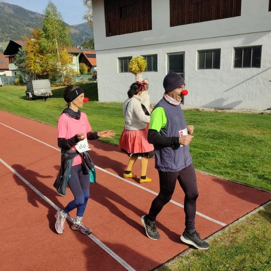 Eine Gruppe von Menschen in Kostümen läuft auf einer Laufbahn, einer trägt eine rote Clownsnase. Die Laufbahn ist von Rasen und Gebäuden umgeben.