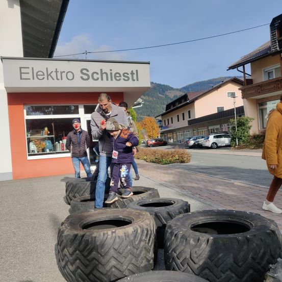 Menschen stehen auf Reifen vor Elektro Schiestl, mit Gebäuden und geparkten Autos dahinter.