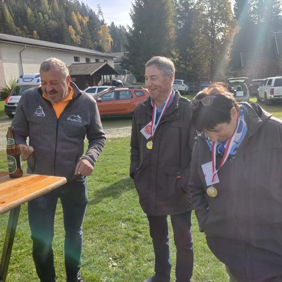 Drei Personen stehen auf einem Rasenfeld und tragen Medaillen. Ein Mann in Grau hält eine Flasche. Hinter ihnen sind Autos geparkt. Bäume und ein Haus sind im Hintergrund.