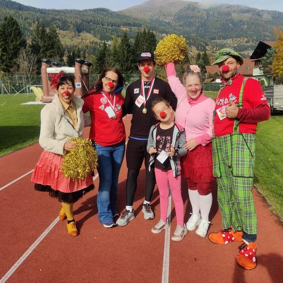 Eine Gruppe von Menschen in Clownkostümen und mit PomPoms posiert für ein Foto auf einer Laufbahn. Im Hintergrund sind Berge und ein grünes Feld zu sehen.