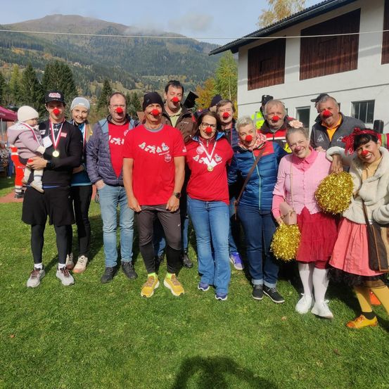 Eine Gruppe von Menschen mit roten Nasen posiert für ein Foto auf einem Rasenfeld vor einem Gebäude mit Bergen im Hintergrund.