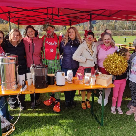 Eine Gruppe von Menschen in Kostümen, darunter Clowns, steht unter einem roten Zelt bei einer Veranstaltung im Freien. Ein Tisch mit Kaffeemaschinen und anderen Gegenständen ist aufgebaut.