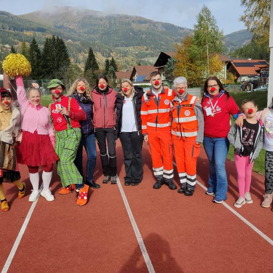Eine Gruppe von Menschen in Clown-Kostümen und mit roten Clownsnasen posiert für ein Foto auf einer Laufbahn mit Bergen im Hintergrund.
